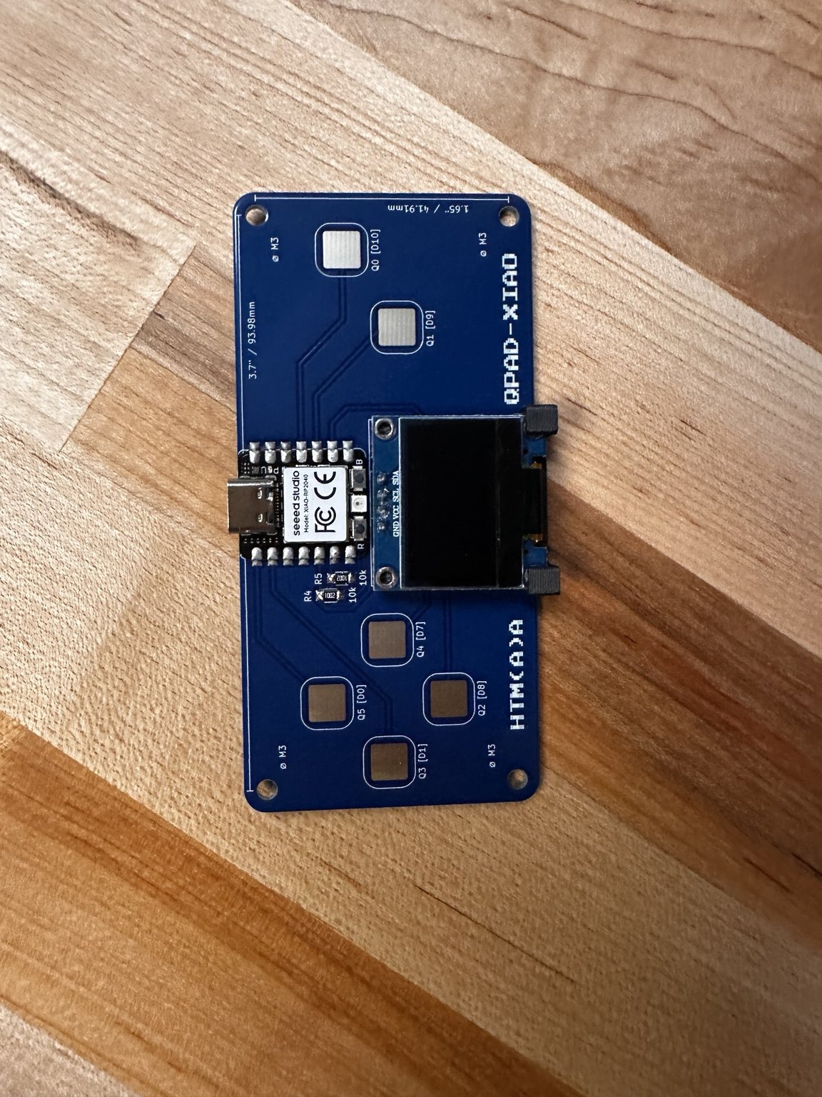 Assembled blue QPAD-XIAO circuit board with OLED and Seeed XIAO RP2040, lying on a wooden table
