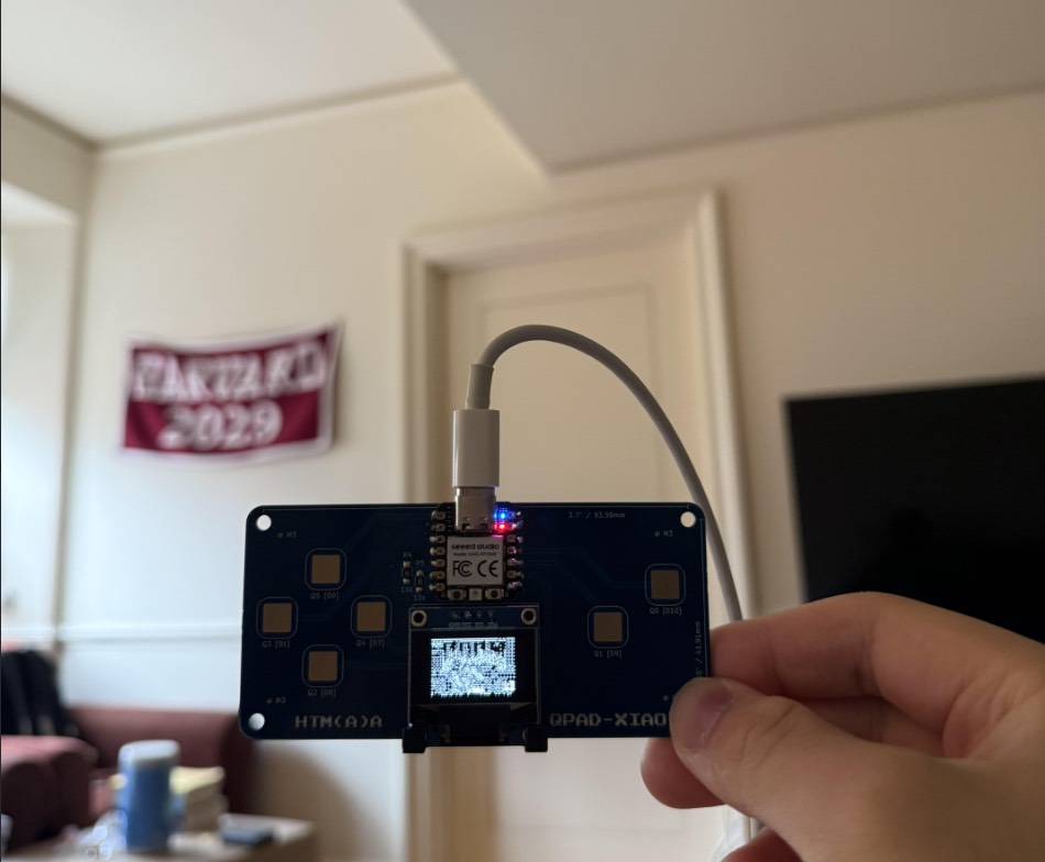 QPAD-XIAO board running Doom on the OLED, held in front of a Harvard 2029 banner