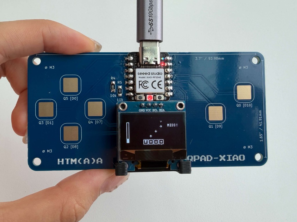 Hand holding a QPAD-XIAO board plugged into USB, with the screen showing a game