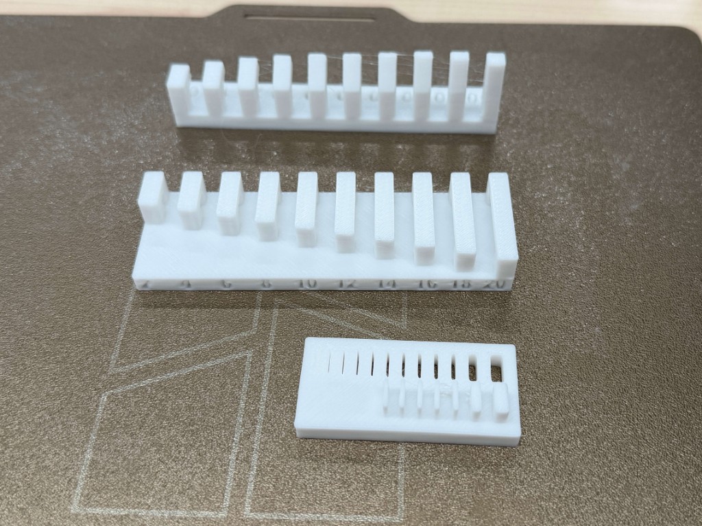 Three white test prints on a printer bed, including a numbered bar and a slot plate
