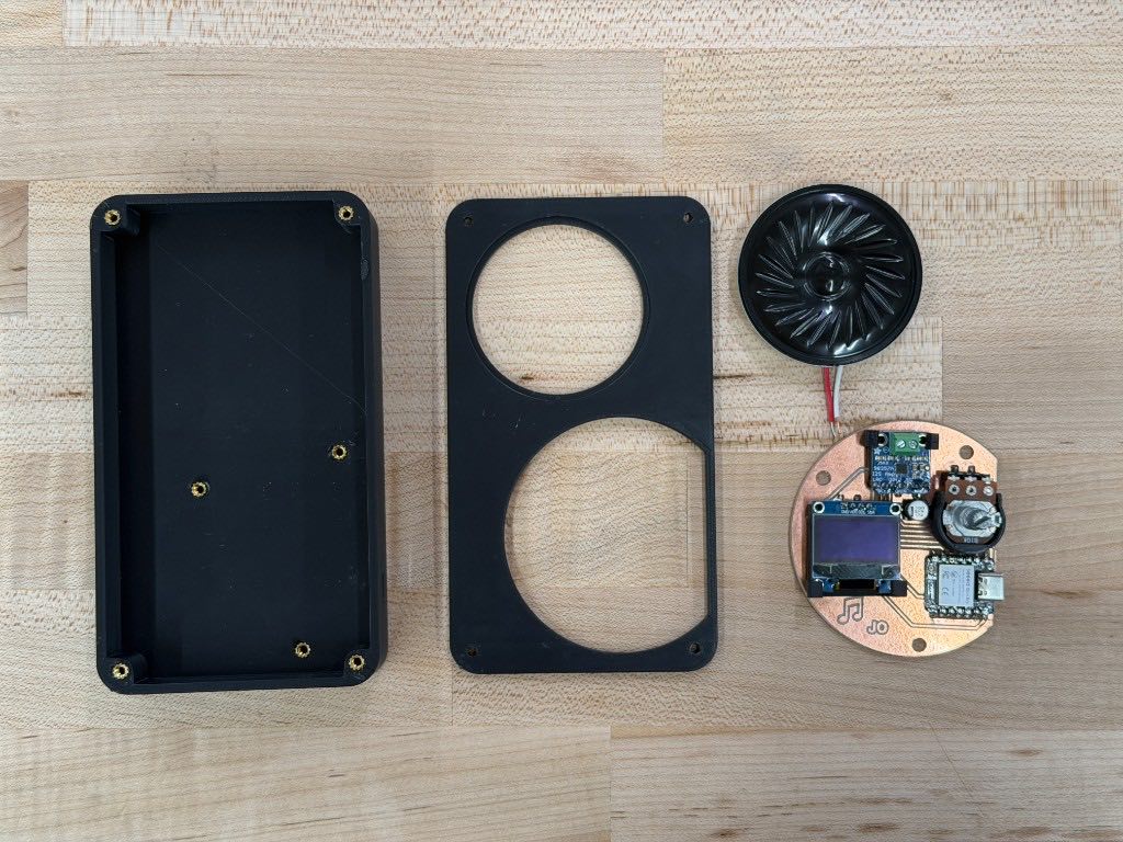 Black enclosure base and lid with two round cutouts, next to the speaker and the assembled circular board with the OLED screen on