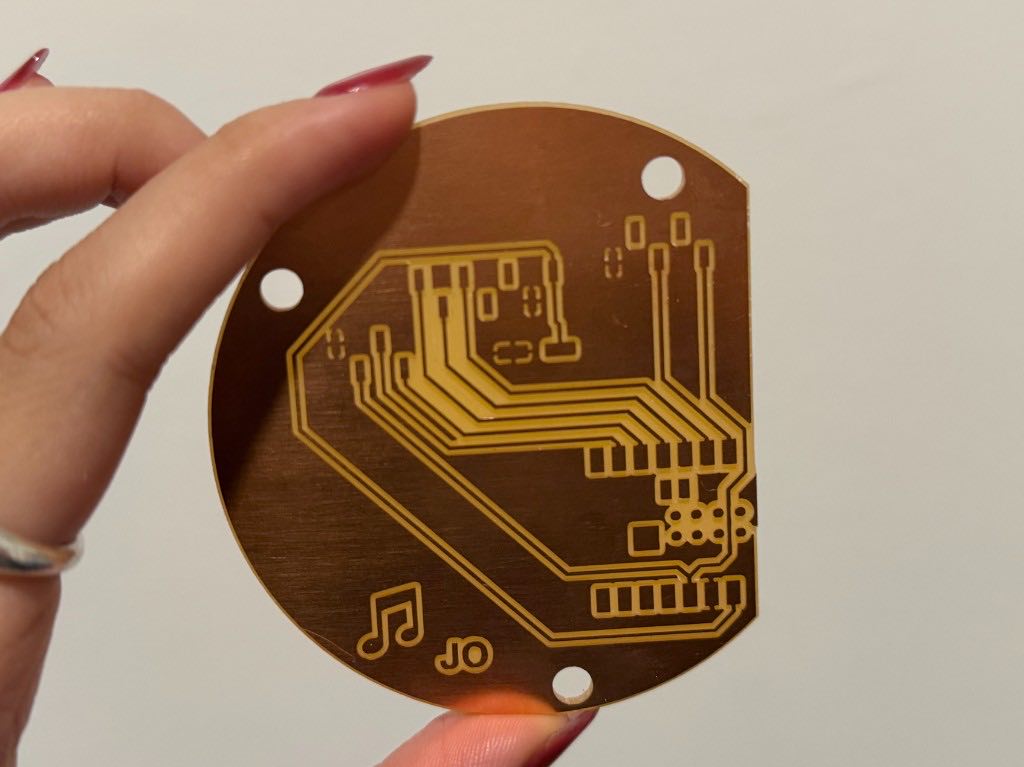 Hand holding the milled circular copper board with traces, mounting holes, a music note, and JO