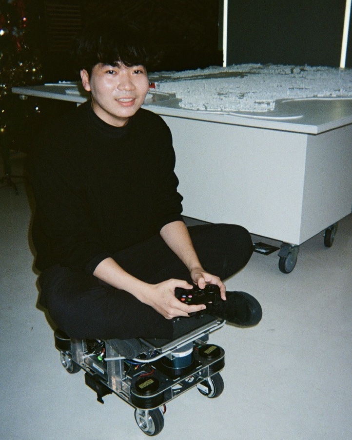 Syuan-Bo sitting cross-legged on a small wheeled robot, holding a game controller, in front of a white city model