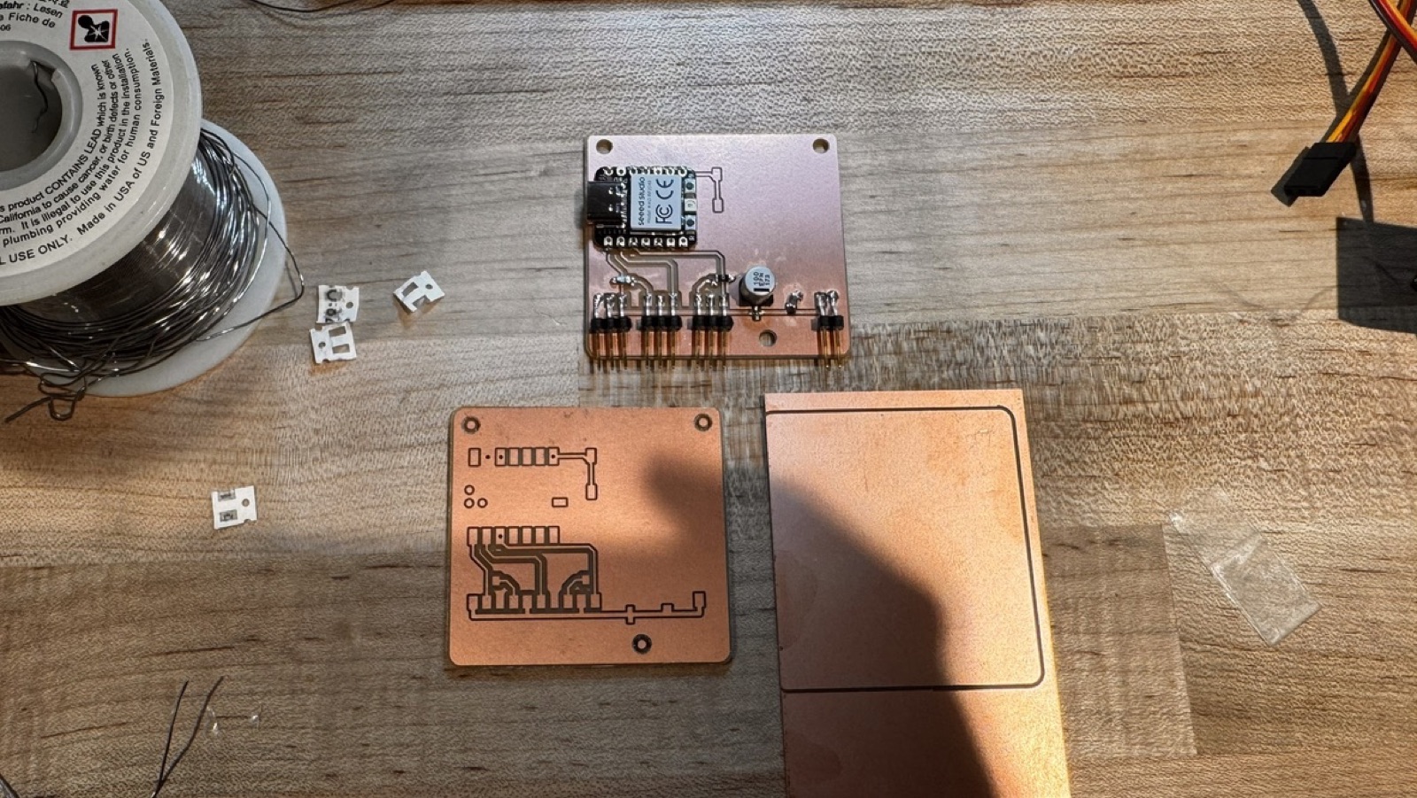 Three boards on a table: the soldered version 1 at the top, an unsoldered milled board at the lower left, and a cut copper blank at the lower right