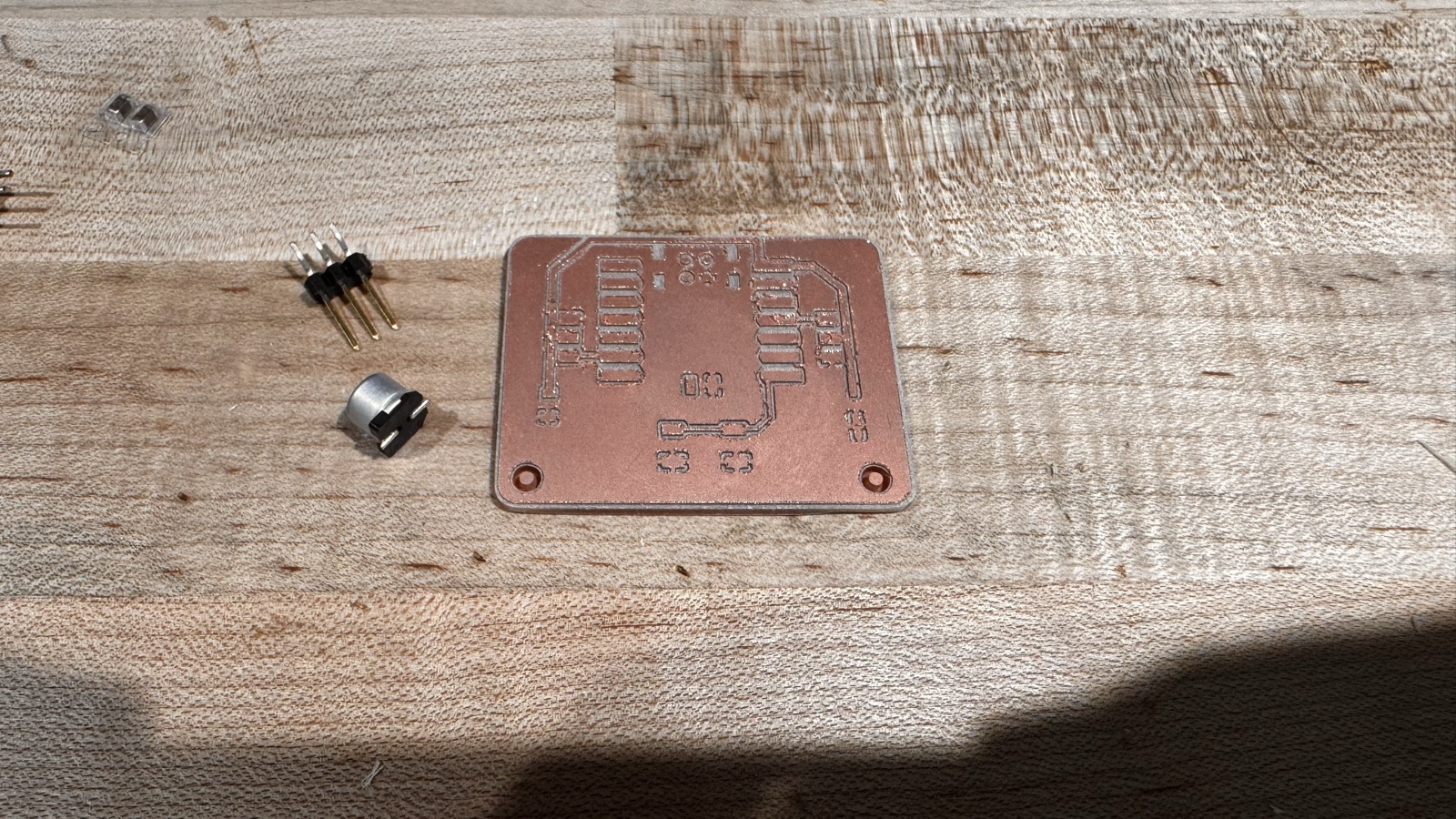 The bare version 2 board on a wooden bench with a pin header and the 100 microfarad capacitor beside it