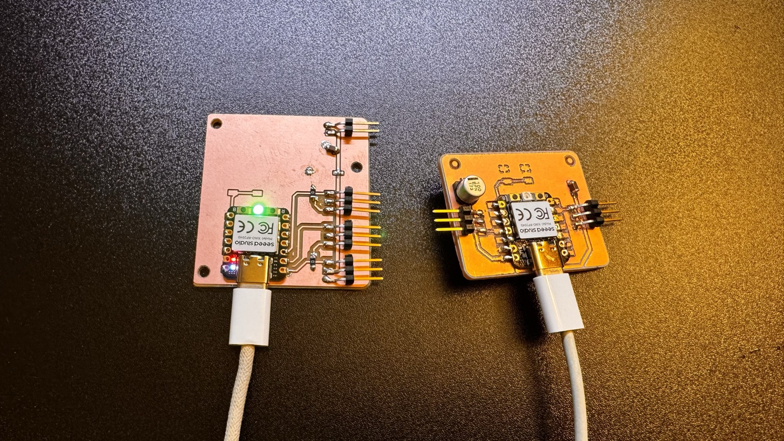 Two boards side by side powered by USB-C: on the left version 1 with a lit green LED on the XIAO, on the right version 2 with an unlit XIAO