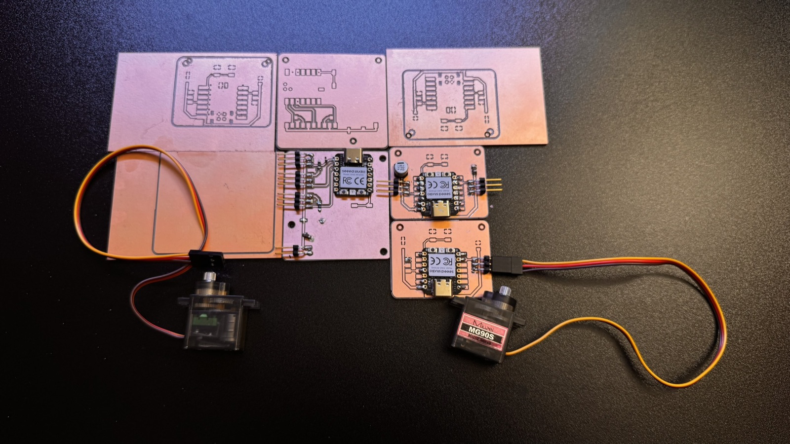 Six milled boards on a black table: three unsoldered boards in the top row, the soldered version 1 in the middle, two soldered version 2 boards at the right, and a servo connected to each of the two boards at the edges