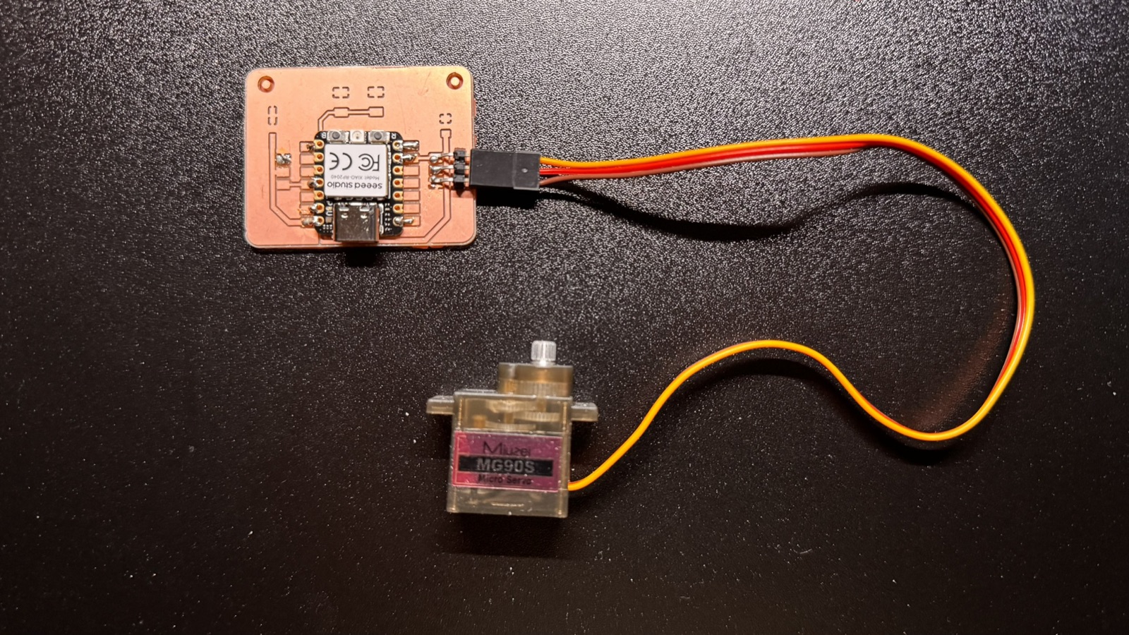The latest version 2 board with a servo plugged into its three-pin header by a cable, lying next to an MG90S servo