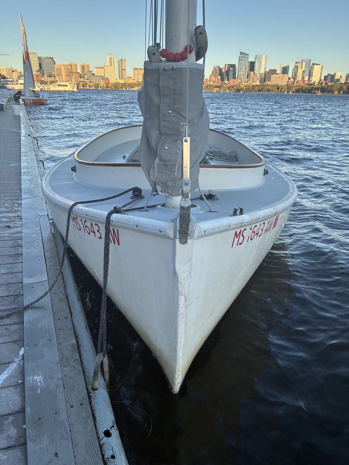Bow view of a white MIT Sailing Pavilion catboat moored at the dock on the Charles River, with its single mast at the very front and the Boston skyline behind