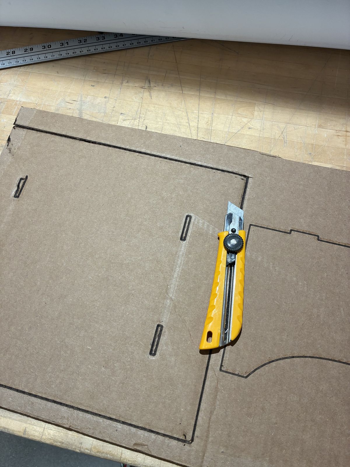A yellow utility knife lying on the laser-engraved cardboard sheet: the rectangular base with two slots on each side, and next to it the outline of one curved support