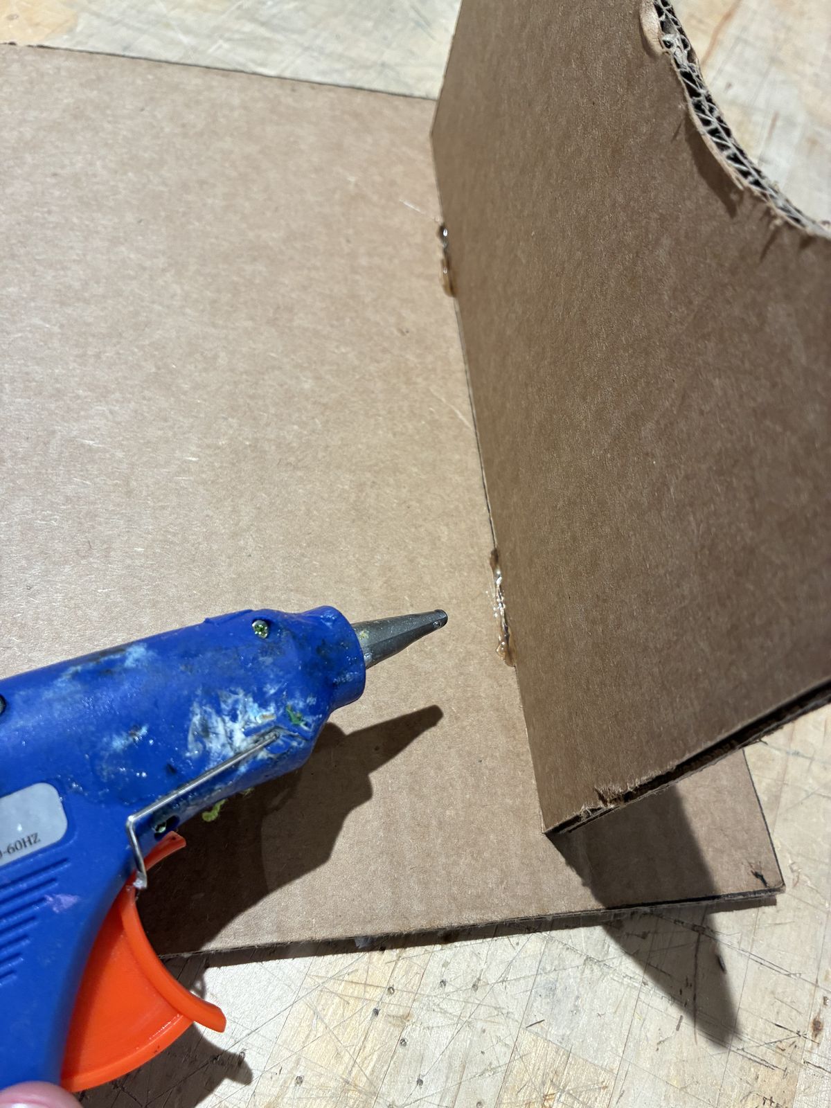 A blue glue gun pointing at the joint between the cardboard base and one of the upright supports, with beads of hot glue along the bottom edge of the support