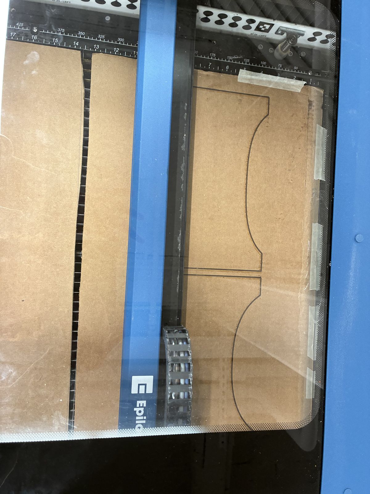 View through the lid of an Epilog laser cutter: a sheet of cardboard taped to the bed, with the outlines of two cradle pieces with curved tops engraved on it but not cut through