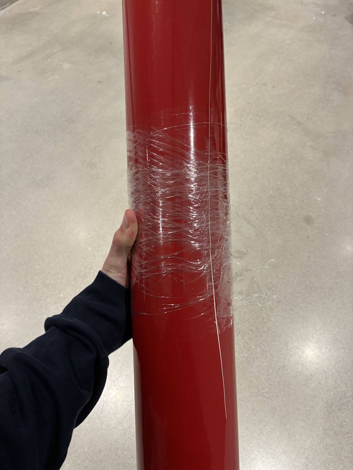 A large roll of glossy red vinyl, wrapped in plastic film, held upright in Victor's hand over a concrete floor