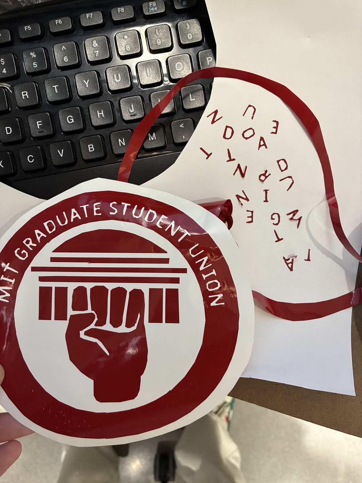 The finished red MIT Graduate Student Union sticker on its backing, with a fist holding up a row of columns, next to the leftover red vinyl and the tiny cut-out letters scattered on a white sheet