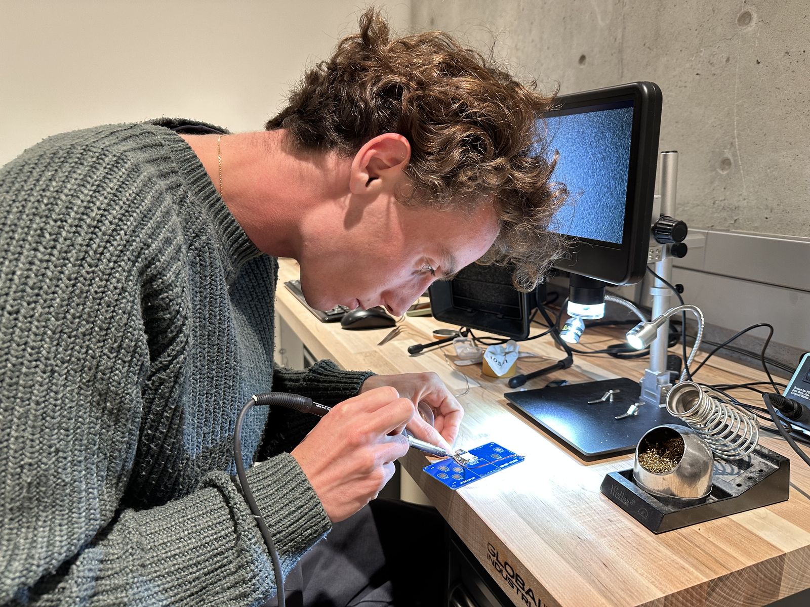 Victor leaning over a workbench with a soldering iron in one hand, working on a small blue circuit board lit by the microscope lamp