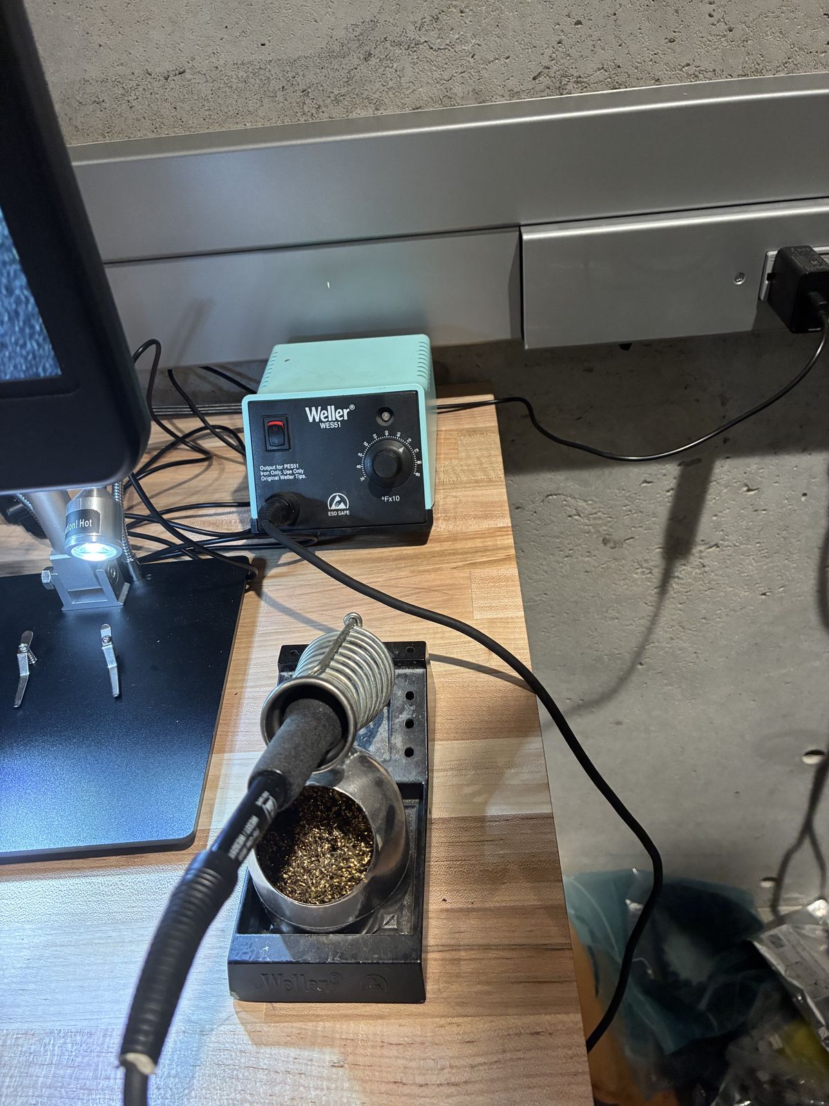 Soldering bench with a light blue Weller WES51 control box, the iron resting in its stand next to a pot of brass wool, and a microscope arm on the left