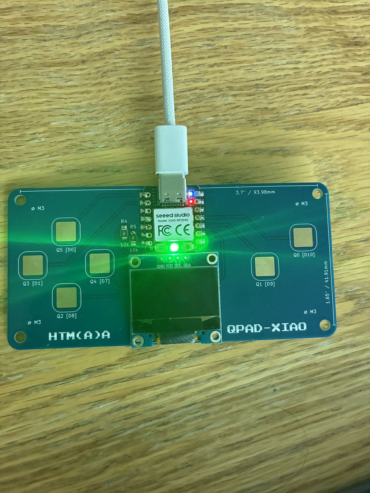 The finished QPAD-XIAO board on a wooden table, plugged into a white USB-C cable, with its green onboard LED lit and the six touch pads and OLED screen visible