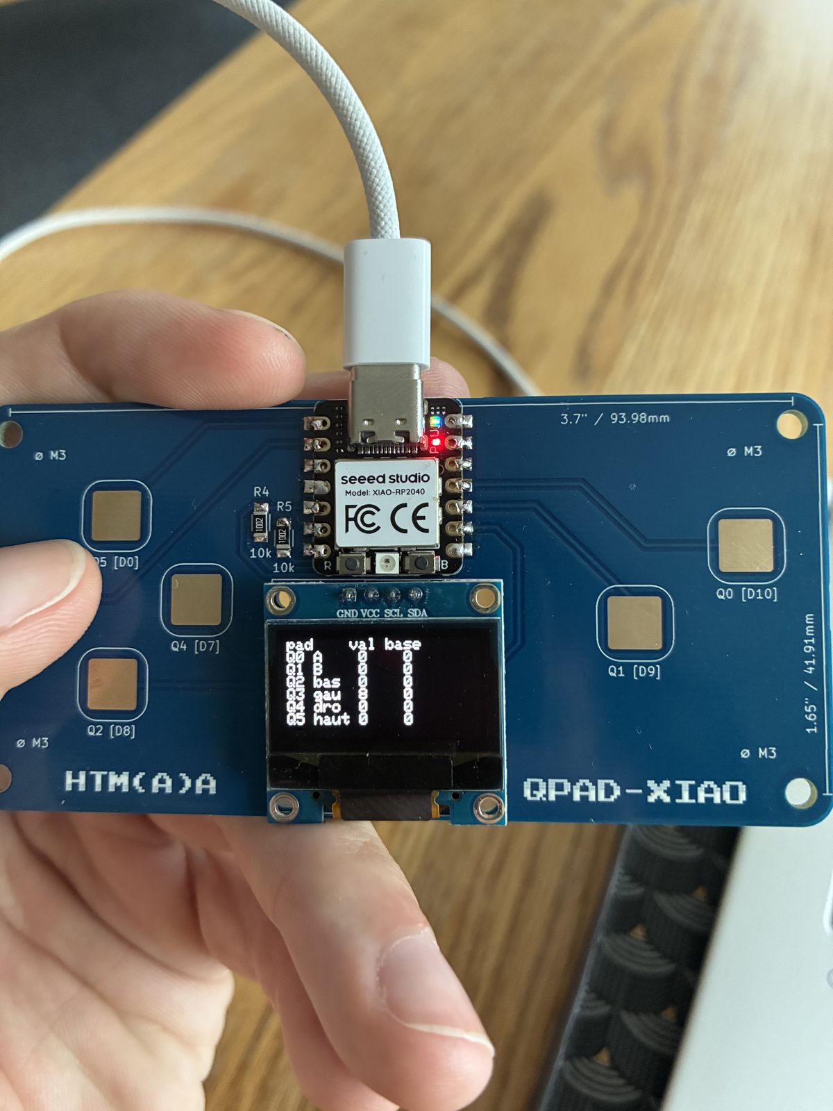 The QPAD-XIAO held in one hand, its OLED showing a table with columns pad, val and base, listing the six pads Q0 to Q5 with their live and baseline values