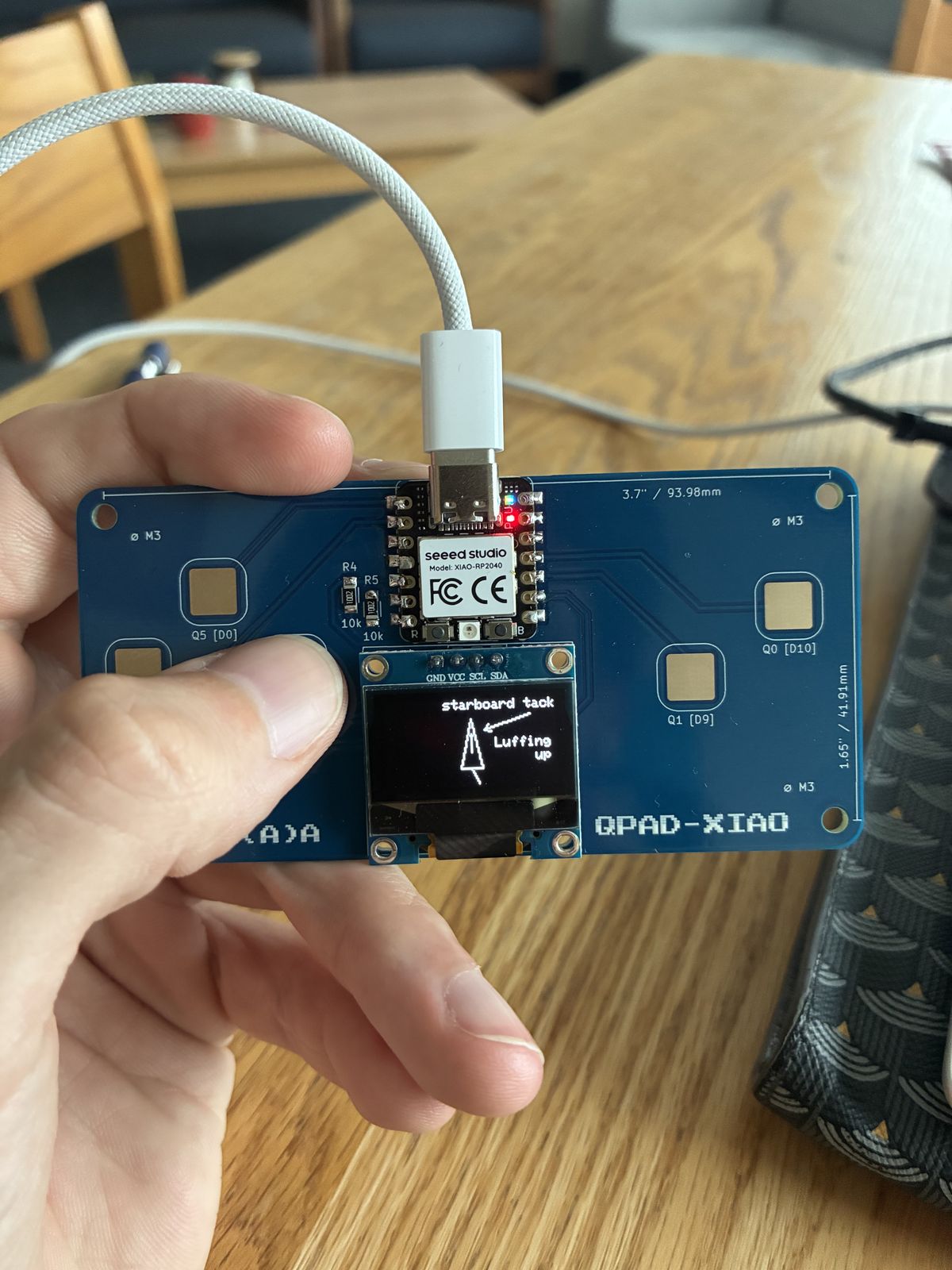 Hand holding the QPAD-XIAO board, its OLED screen showing the words starboard tack at the top, an arrow pointing down at a triangular boat, and the message Luffing up on the right