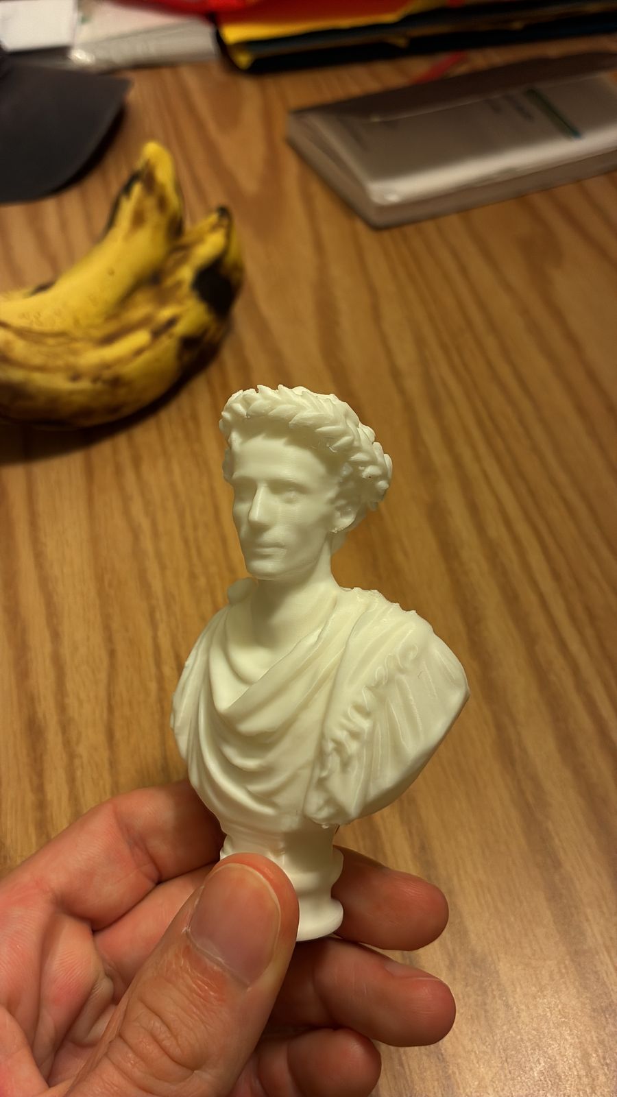 The finished white 3D-printed bust held in Victor's hand: his own face with a laurel wreath, on a draped Roman toga and a round pedestal, with bananas on the table behind