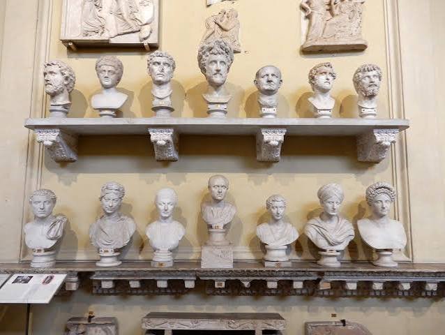 Two long marble shelves in the Vatican Museums lined with fourteen Roman portrait busts of men and women, with carved reliefs on the wall above