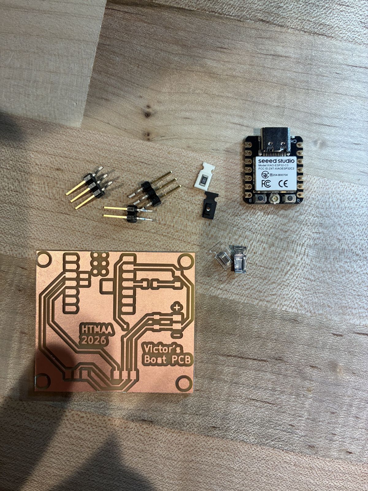 The bare milled board on a wooden bench next to all its parts: the XIAO ESP32-C3 module, two 3-pin and one 2-pin right-angle headers, a resistor marked 1001 on its white tape, and the other tiny surface-mount parts still in their packaging
