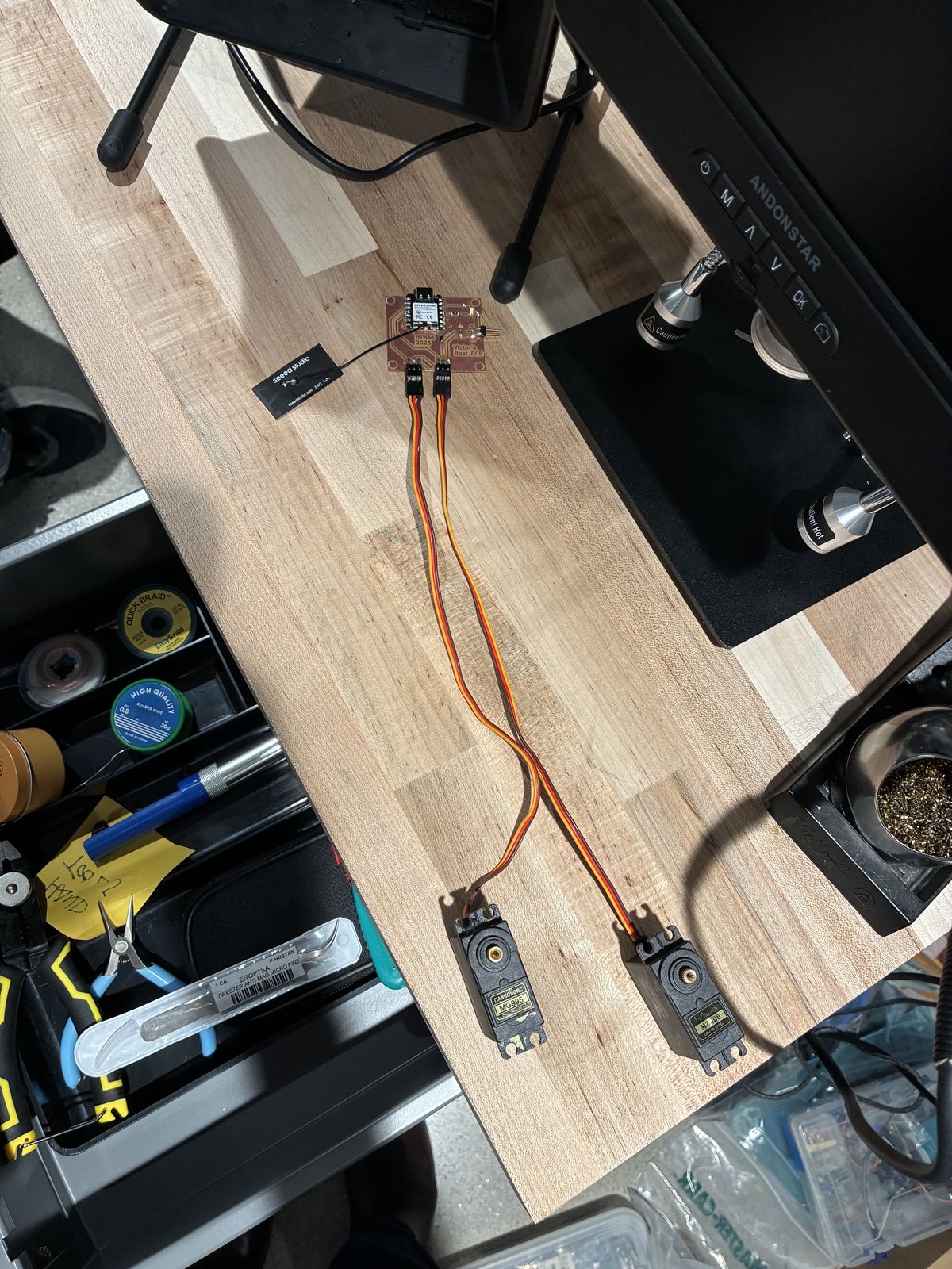 View from above of a workbench: the finished board with two servo cables plugged into its headers, leading to two black servos