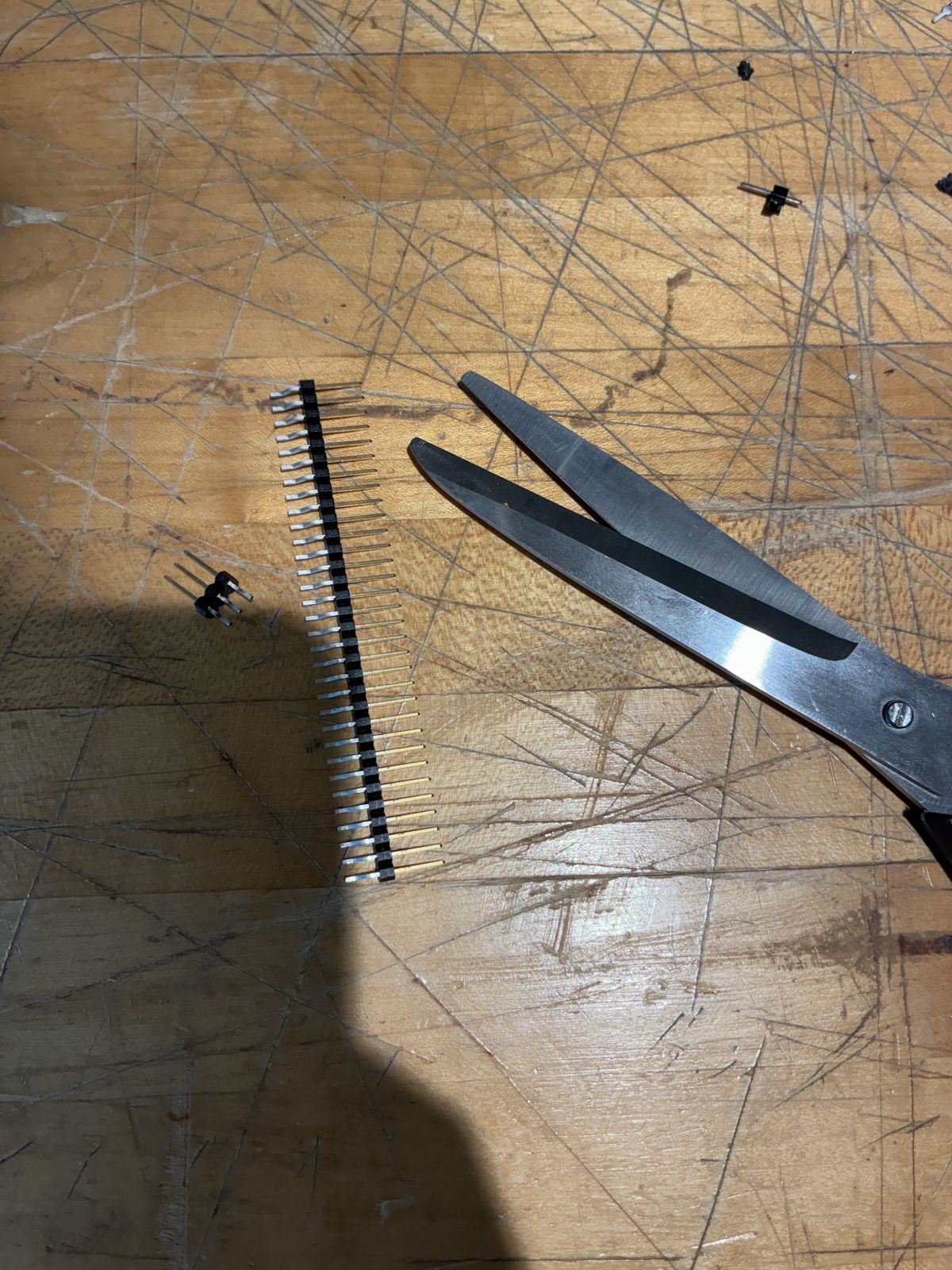 A long strip of right-angle header pins lying on a scratched wooden workbench next to a pair of scissors, with a short 2-pin piece already cut off