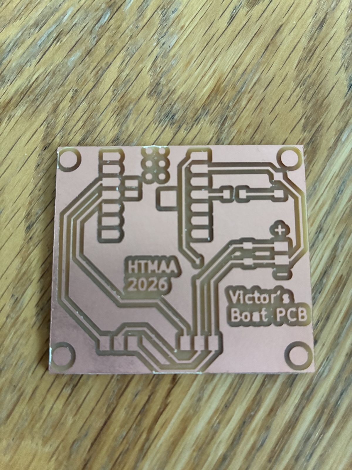 The bare milled copper board on a wooden table, cut out and sanded, showing the XIAO pads, the tracks, the plus and minus signs, the text HTMAA 2026 and Victor's Boat PCB, and four M3 holes in the corners
