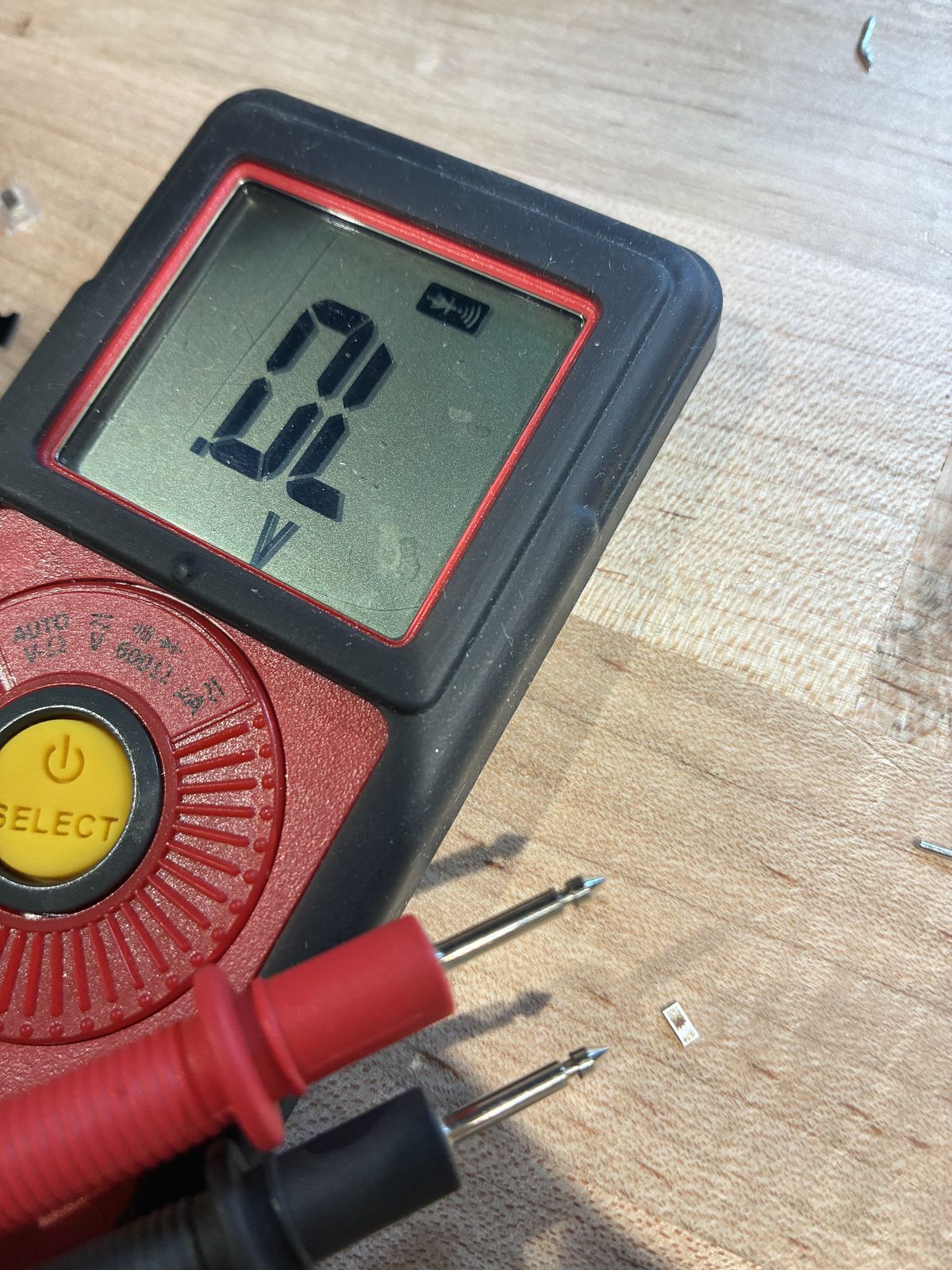 A red and black multimeter set to diode test mode showing OL on its screen, its two probes lying next to a tiny surface-mount LED on the wooden bench