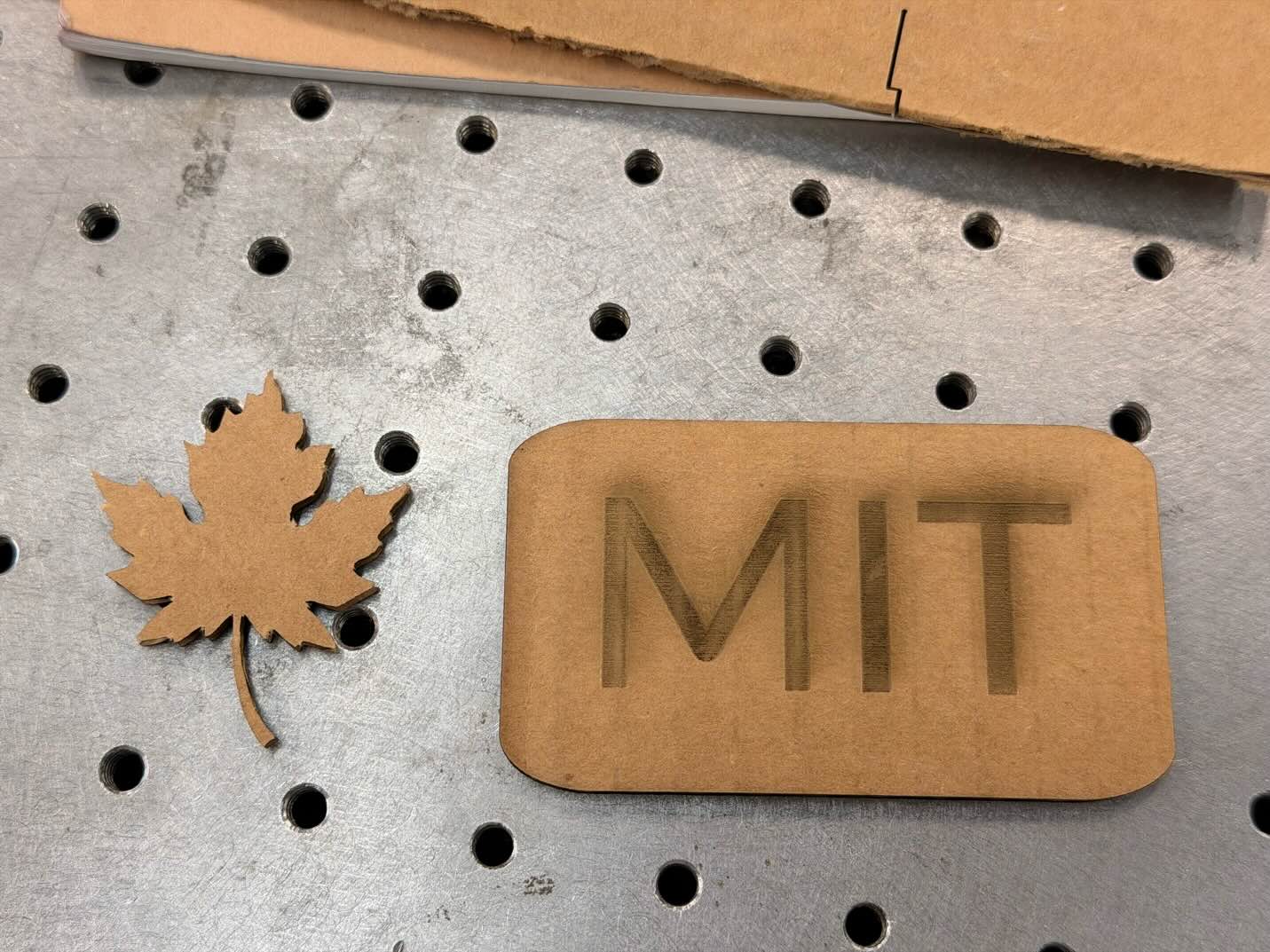 A maple-leaf cutout beside an MIT engraving on the laser cutter bed