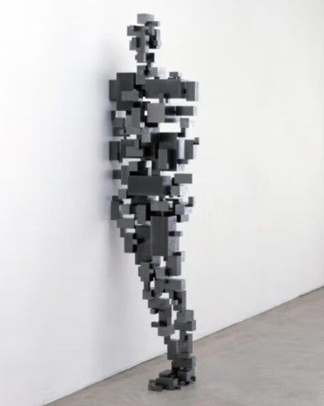 A figure by Antony Gormley made of stacked dark blocks, leaning on a wall